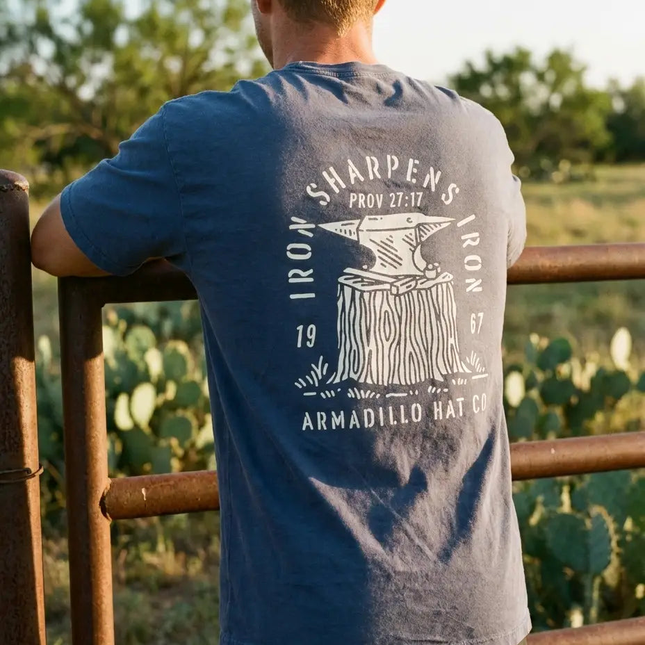 Man in a ball cap leaning on a rusted pipe ranch gate wearing the Forged Tee, Iron Sharpens Iron back print, prickly pear in background