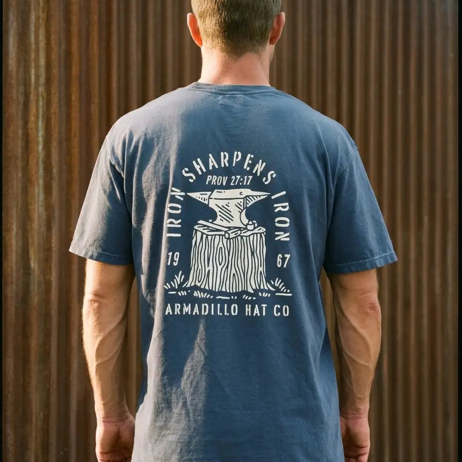 Forged Tee back print, cream anvil on stump with Iron Sharpens Iron Prov 27:17, on washed navy heavyweight tee against rusted steel