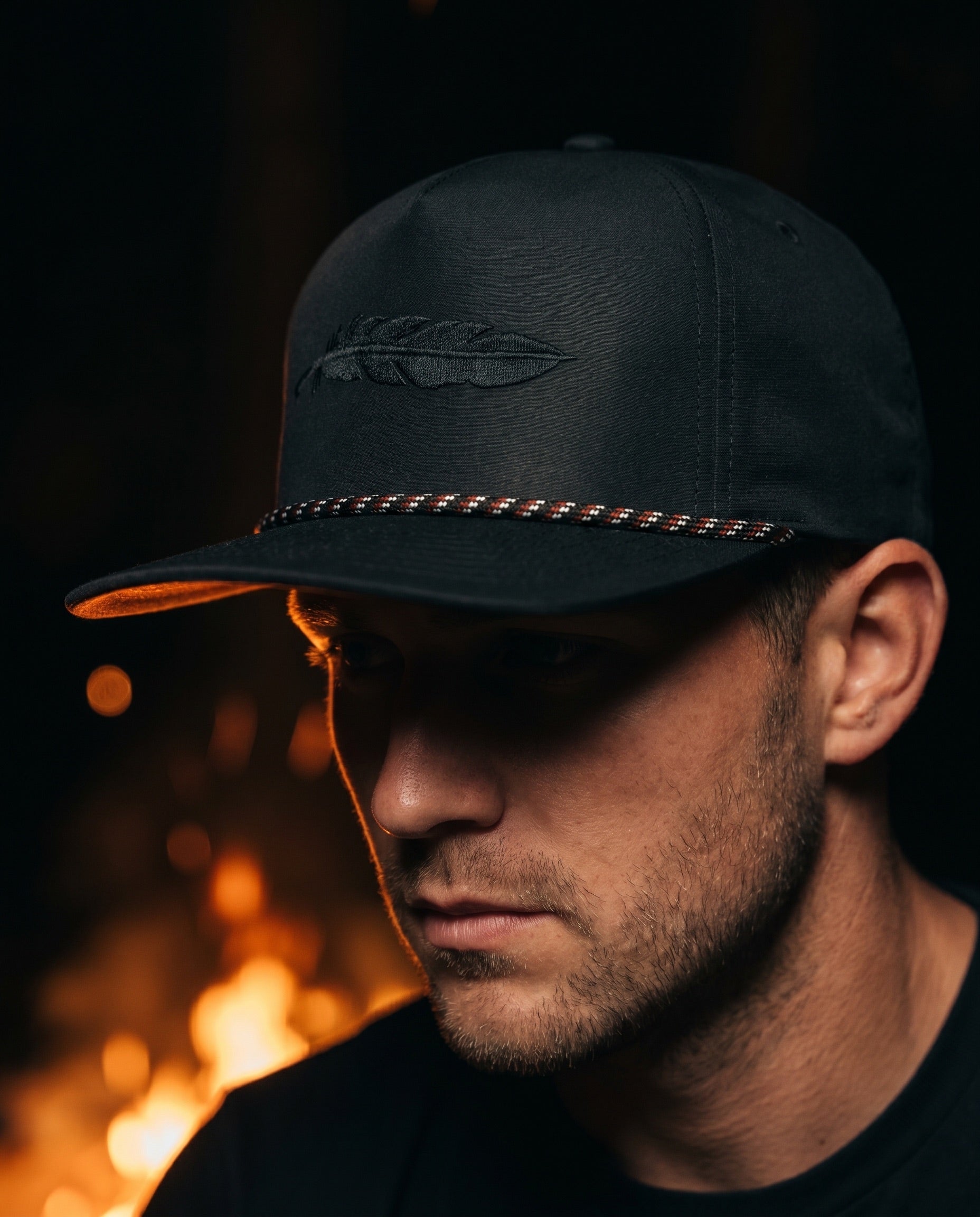 Man in a black Armadillo Hat Co. rope snapback lit by a campfire at night
