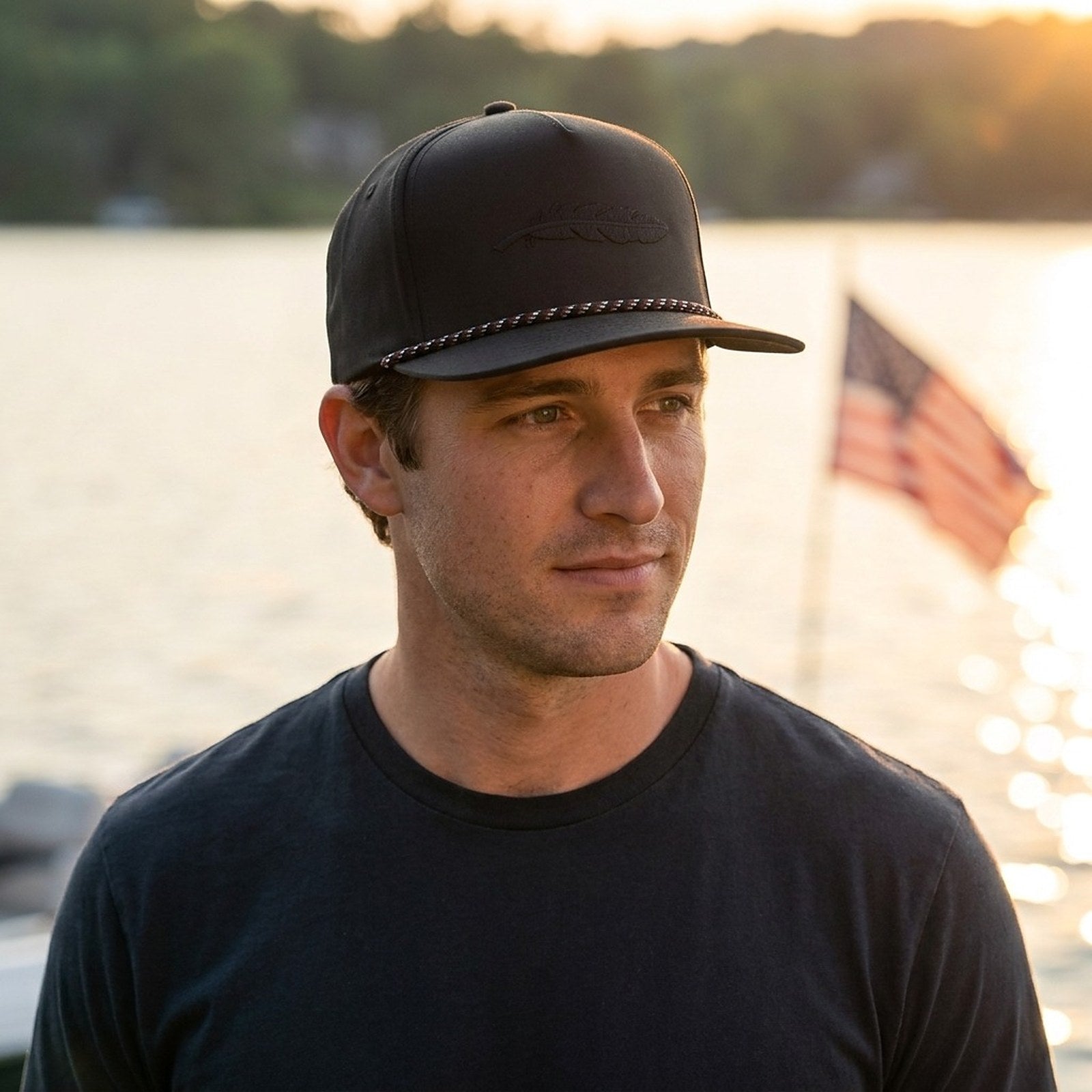 Man in a black Armadillo Hat Co. rope snapback by the lake at sunset with an American flag flying behind him