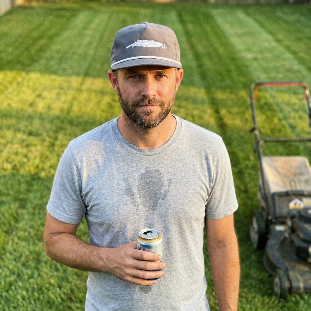 Man in the grey Scrambler rope cap with the white AHC feather, cold drink in hand after mowing, striped lawn and mower behind