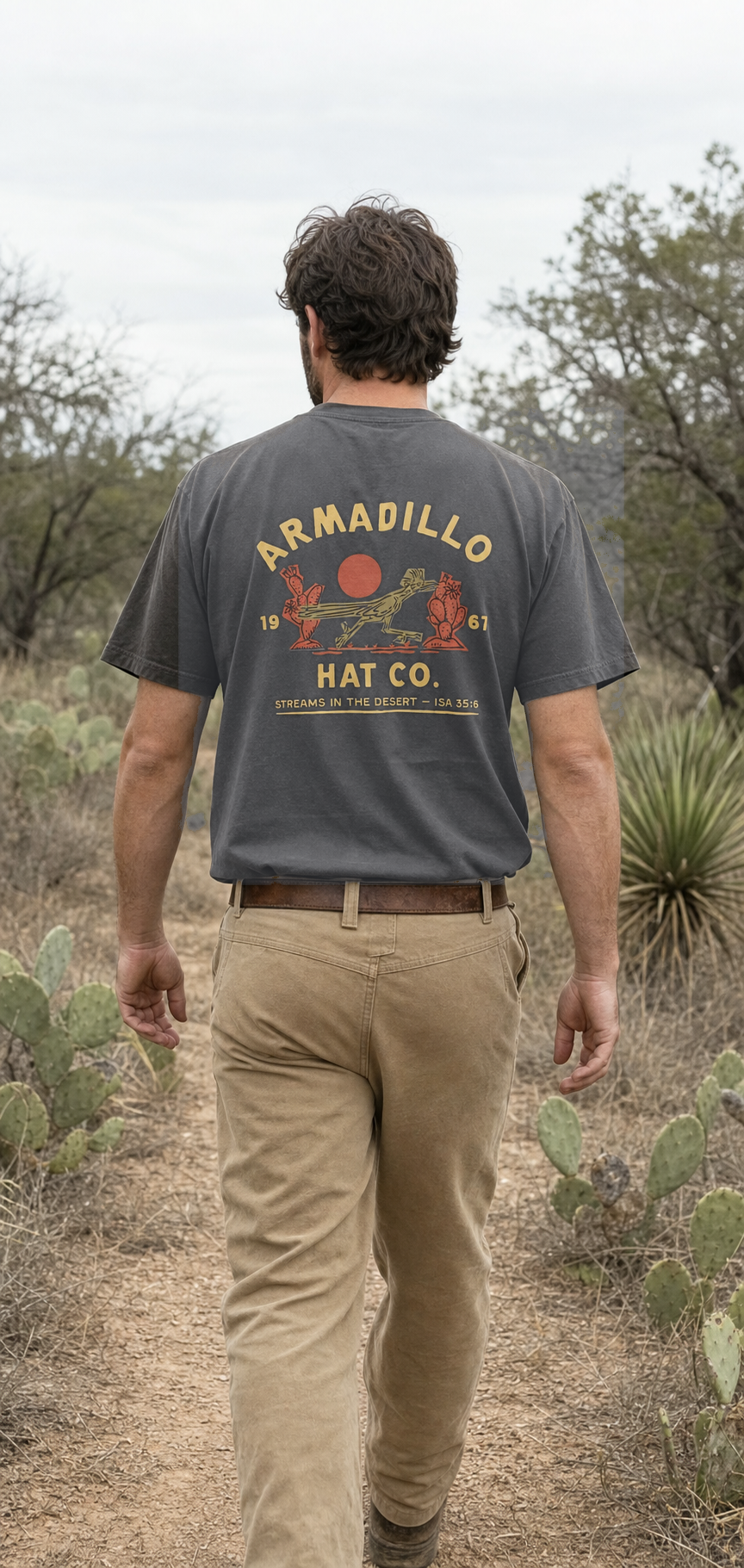 Man walking a desert two-track through prickly pear wearing the Roadrunner Tee in Pepper charcoal, back print with the roadrunner and red sun, Isaiah 35:6