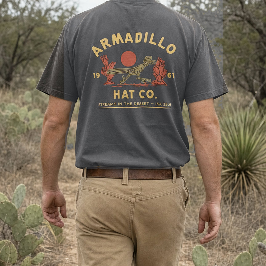 Man walking a desert two-track through prickly pear wearing the Roadrunner Tee in Pepper charcoal, back print with the roadrunner and red sun, Isaiah 35:6