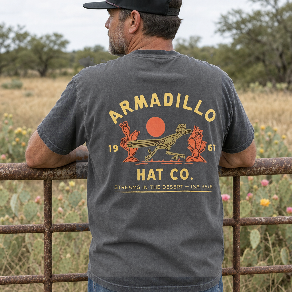 Man in an Armadillo Hat Co rope cap leaning on a rusted ranch gate wearing the Roadrunner Tee in Pepper charcoal, back print with the roadrunner and red sun, Streams in the Desert, Isaiah 35:6