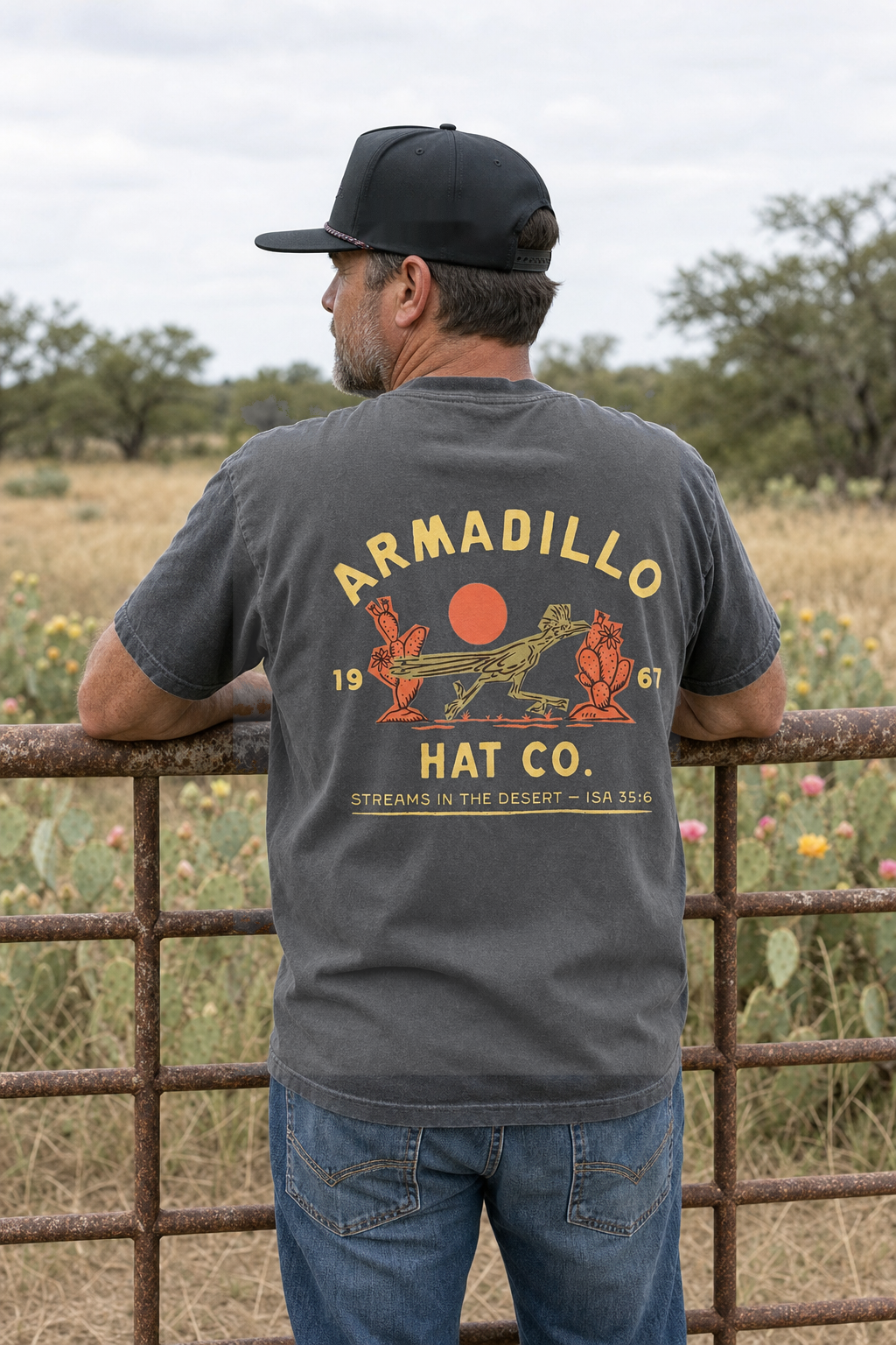 Man in an Armadillo Hat Co rope cap leaning on a rusted ranch gate wearing the Roadrunner Tee in Pepper charcoal, back print with the roadrunner and red sun, Streams in the Desert, Isaiah 35:6