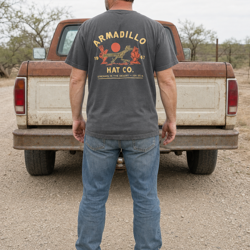 Man standing behind a weathered pickup truck wearing the Roadrunner Tee in Pepper charcoal, full back print visible with Streams in the Desert, Isaiah 35:6